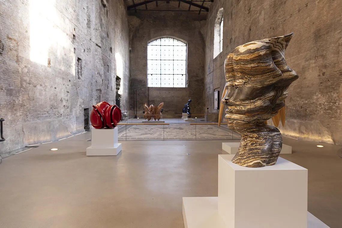 veduta della mostra all'interno delle Terme di Diocleziano con un grande mosaico a pavimento circondato da sculture di materiali e forme diverse di Tony Cragg